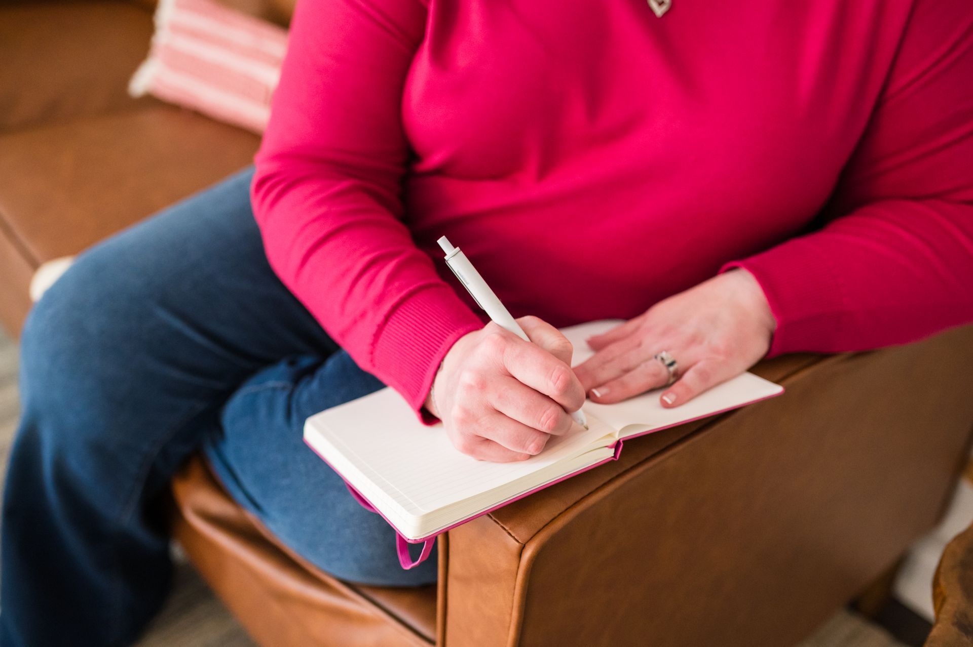 Woman in red top and jeans writes in notebook, sitting on a brown leather couch.