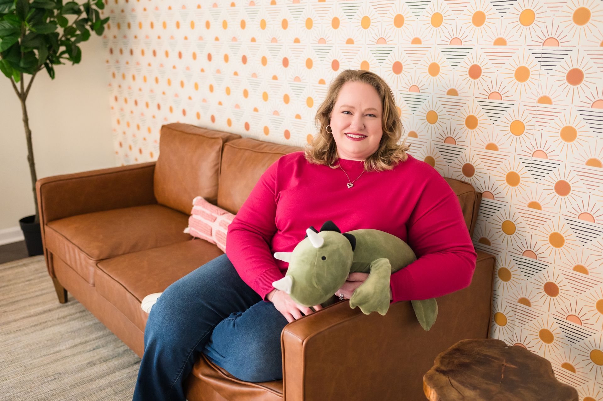 Woman in red shirt and jeans sits on a brown couch, holding a plush dinosaur. Colorful wallpaper.