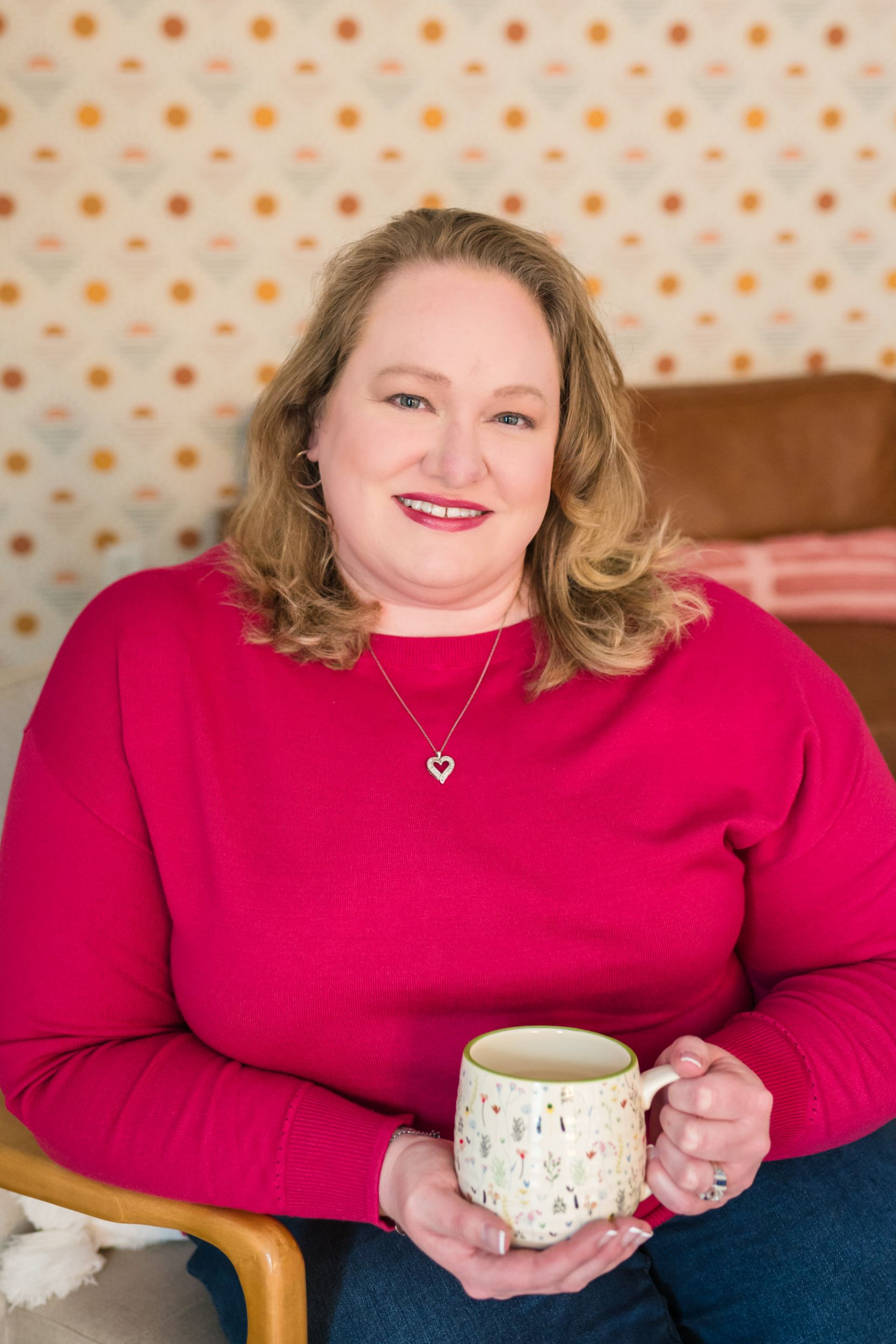 Woman with blonde hair, red sweater, holding a mug, smiles, sitting in a chair, patterned wall background.