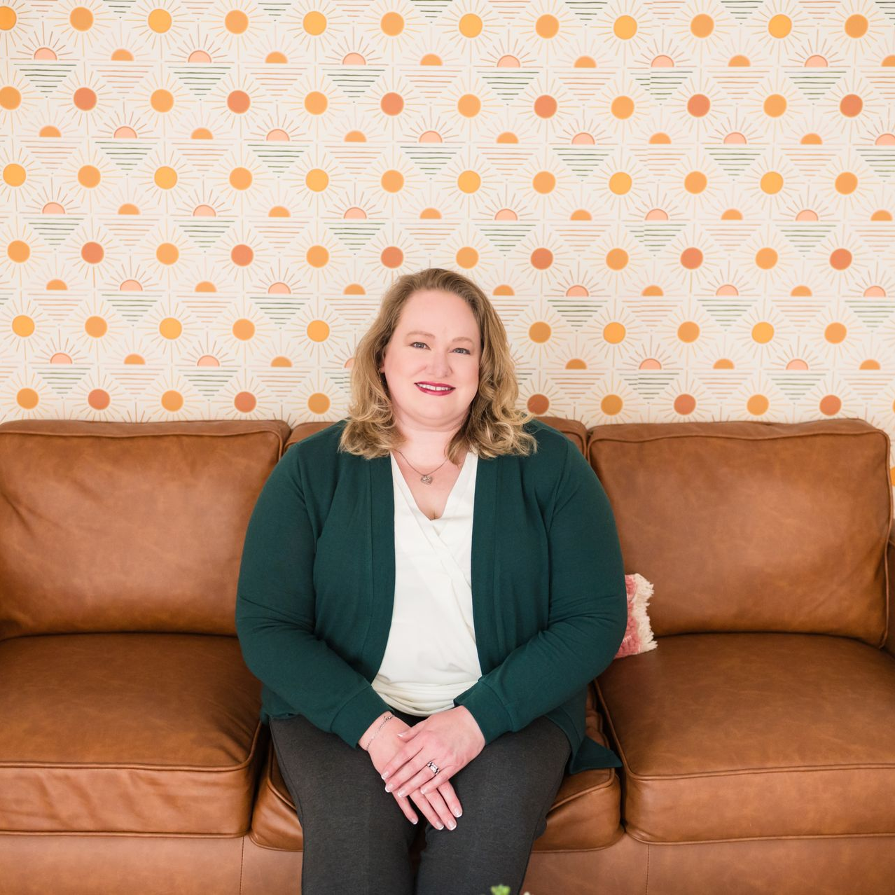 Woman with blonde hair, in green cardigan, sitting on brown couch. Patterned wallpaper backdrop.