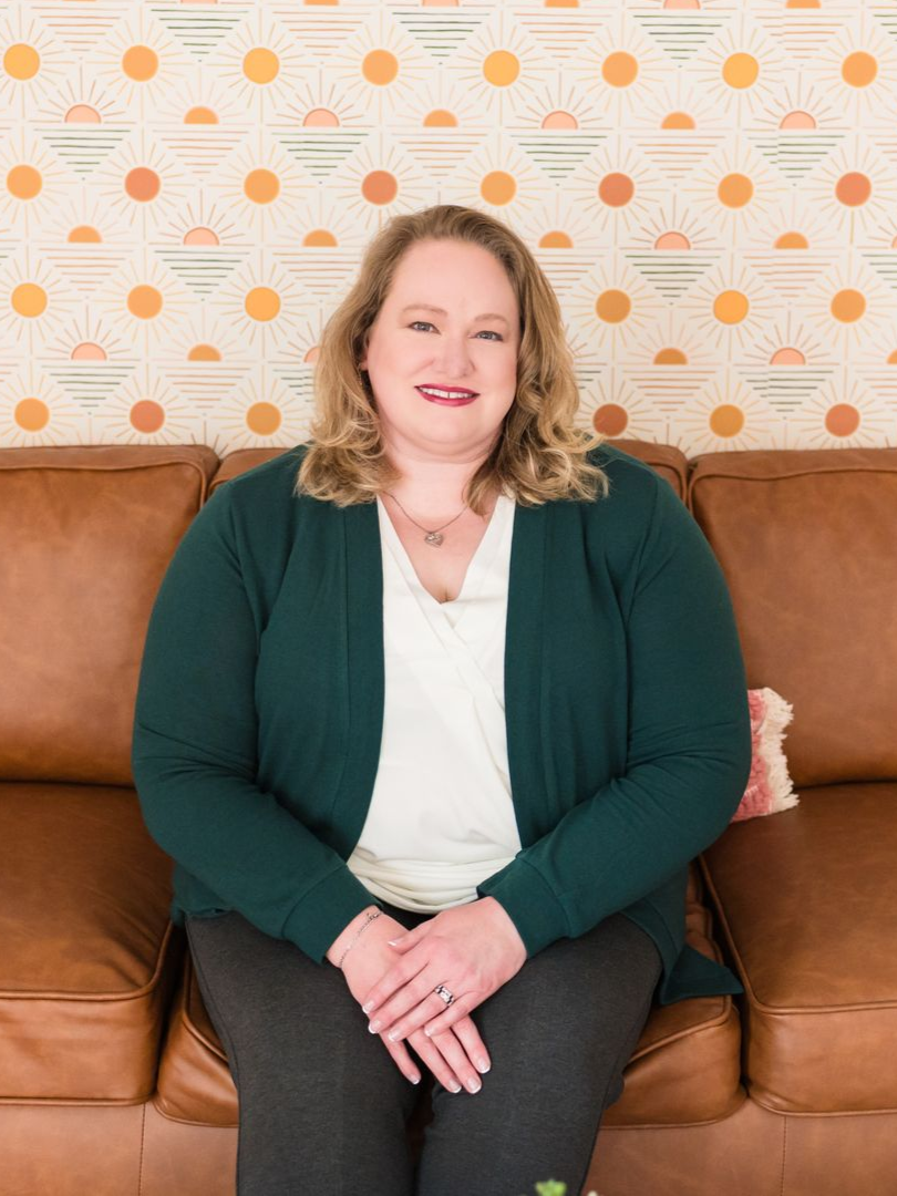 Woman in green cardigan gestures during a video call on a laptop at a wooden desk, home office setting.