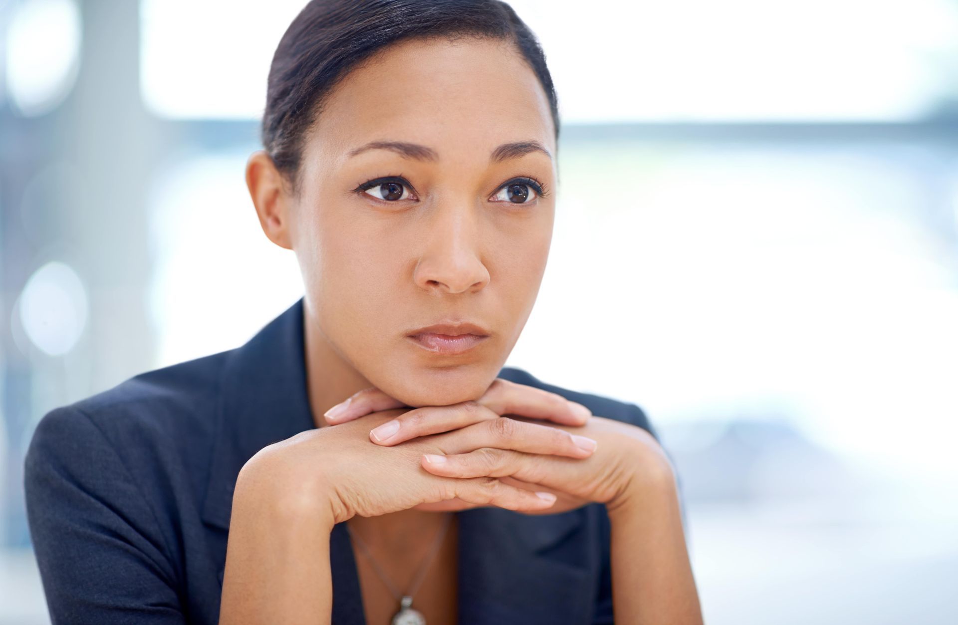 Woman in a navy blazer rests chin on hands, looking thoughtful in a bright office setting.
