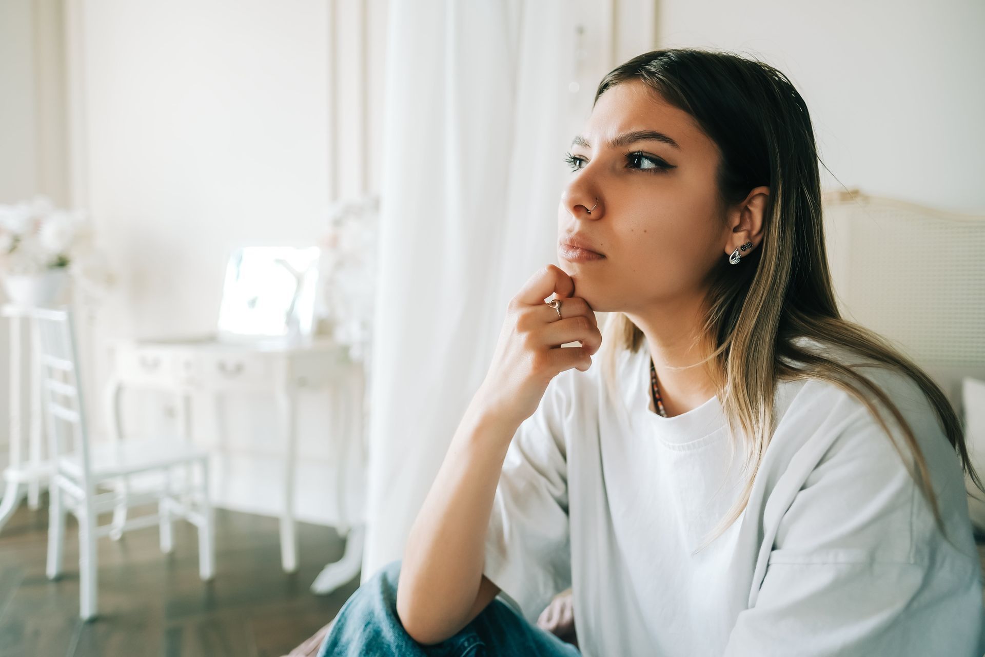 Young woman in white shirt, looking thoughtful, resting chin on hand in a bright room.