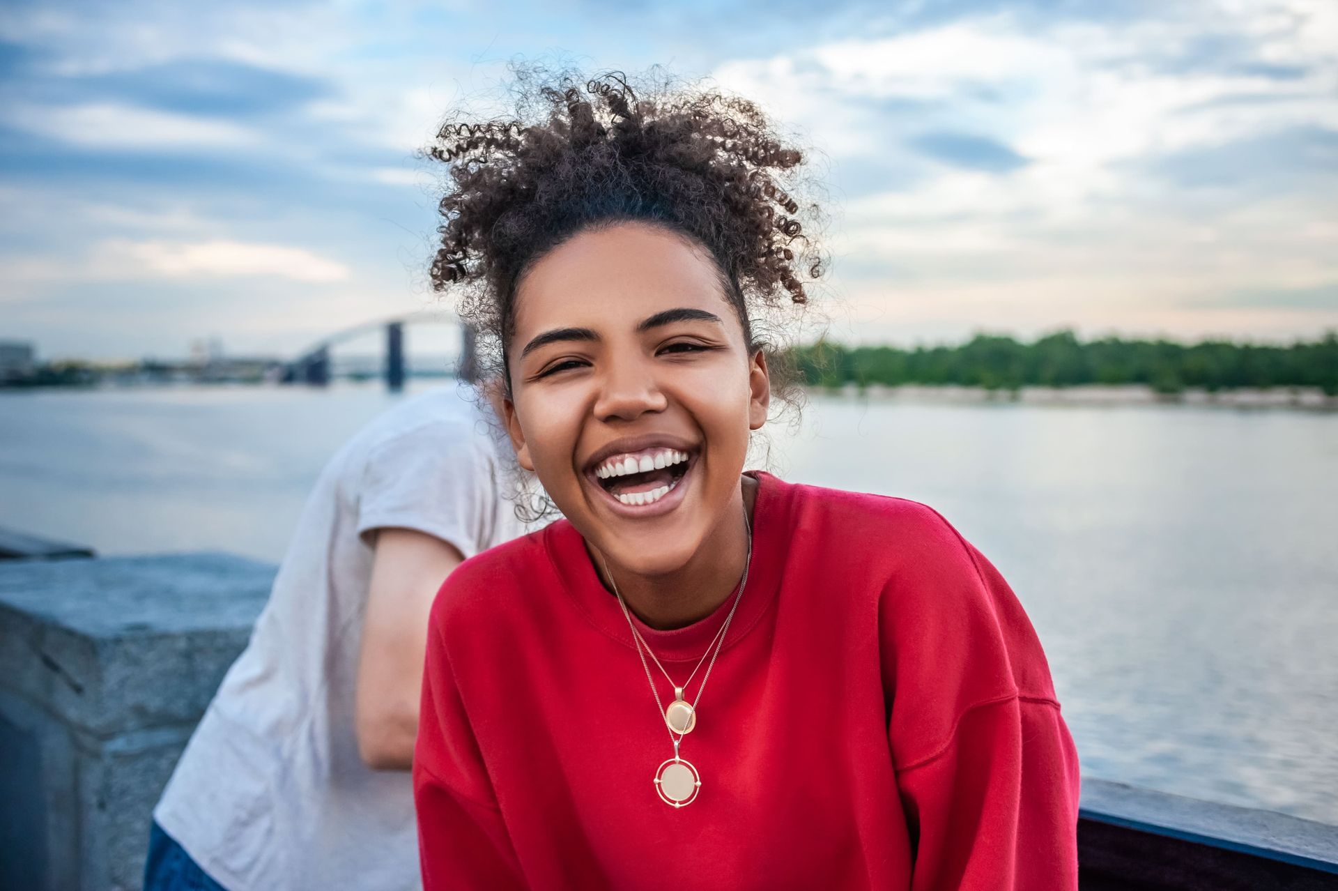 Young Black woman laughs, red sweater, water, and bridge in background.
