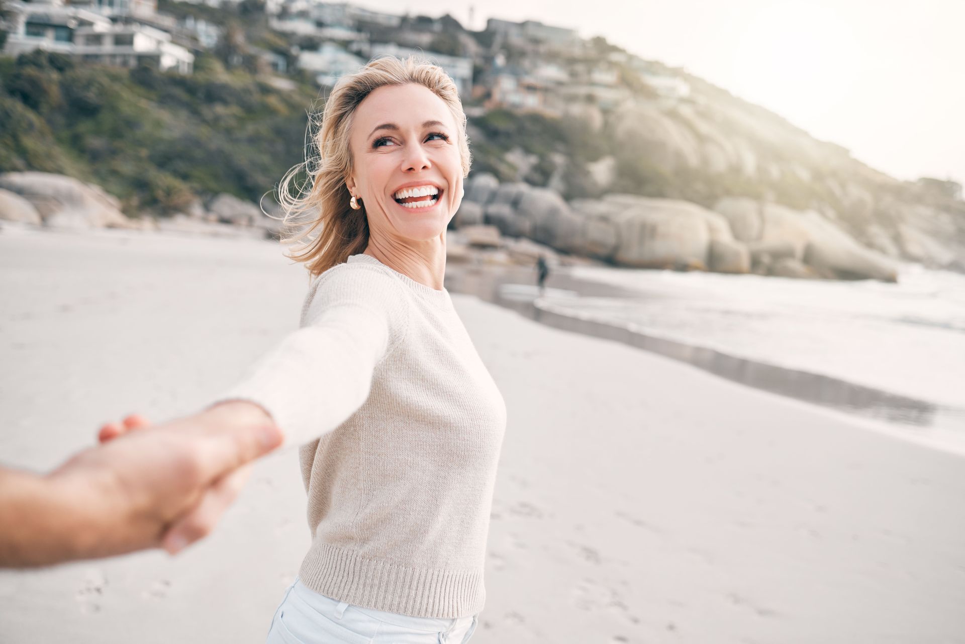 Woman smiles, holding hand on beach, looking toward sunlit cliffs.