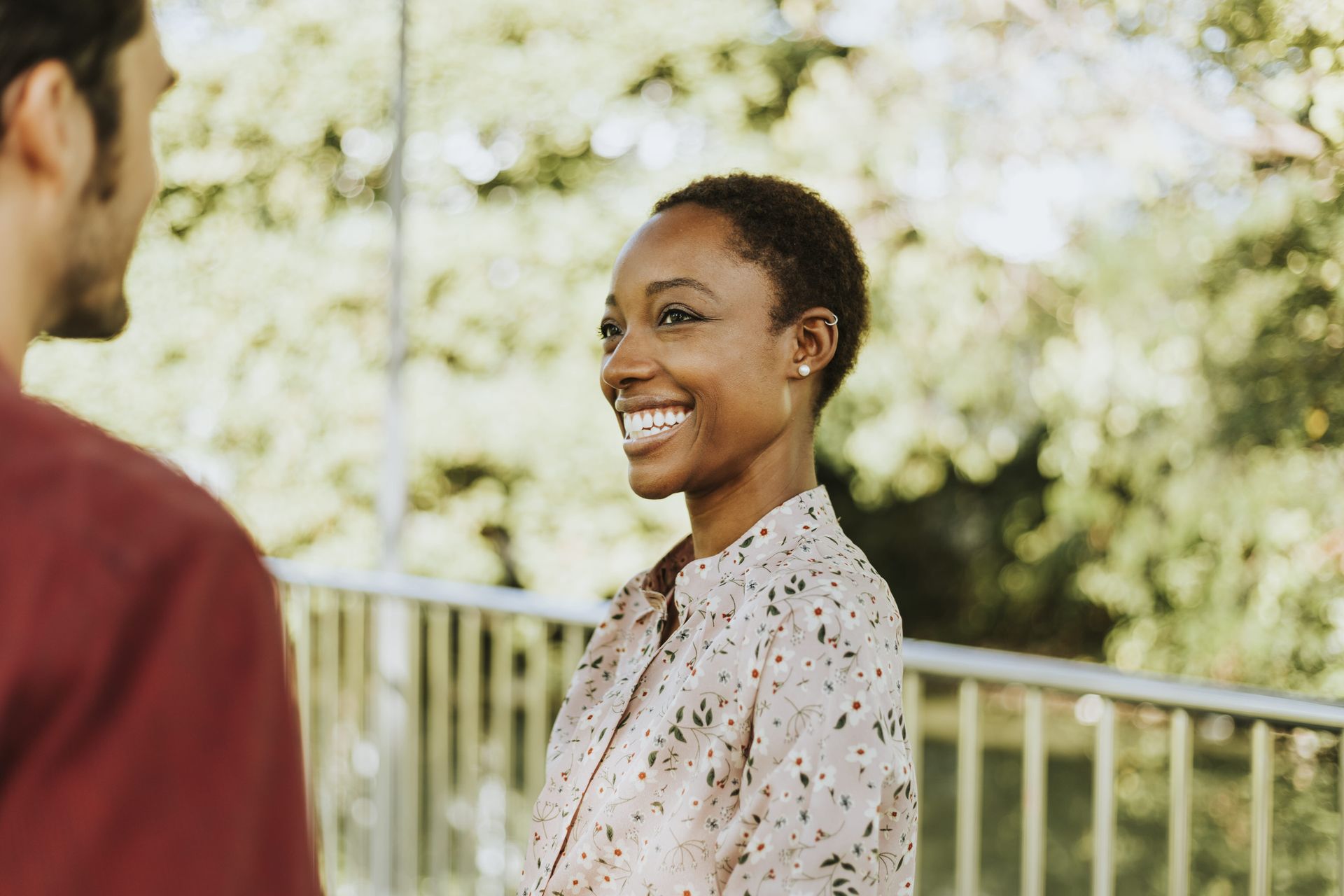 Woman with short hair smiling at a person, outside by a railing and trees.