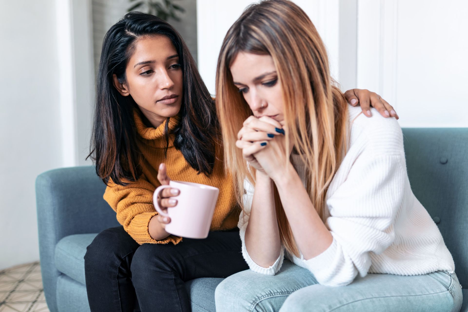 Woman comforts another, offering a shoulder and holding a mug, indoors on a sofa.