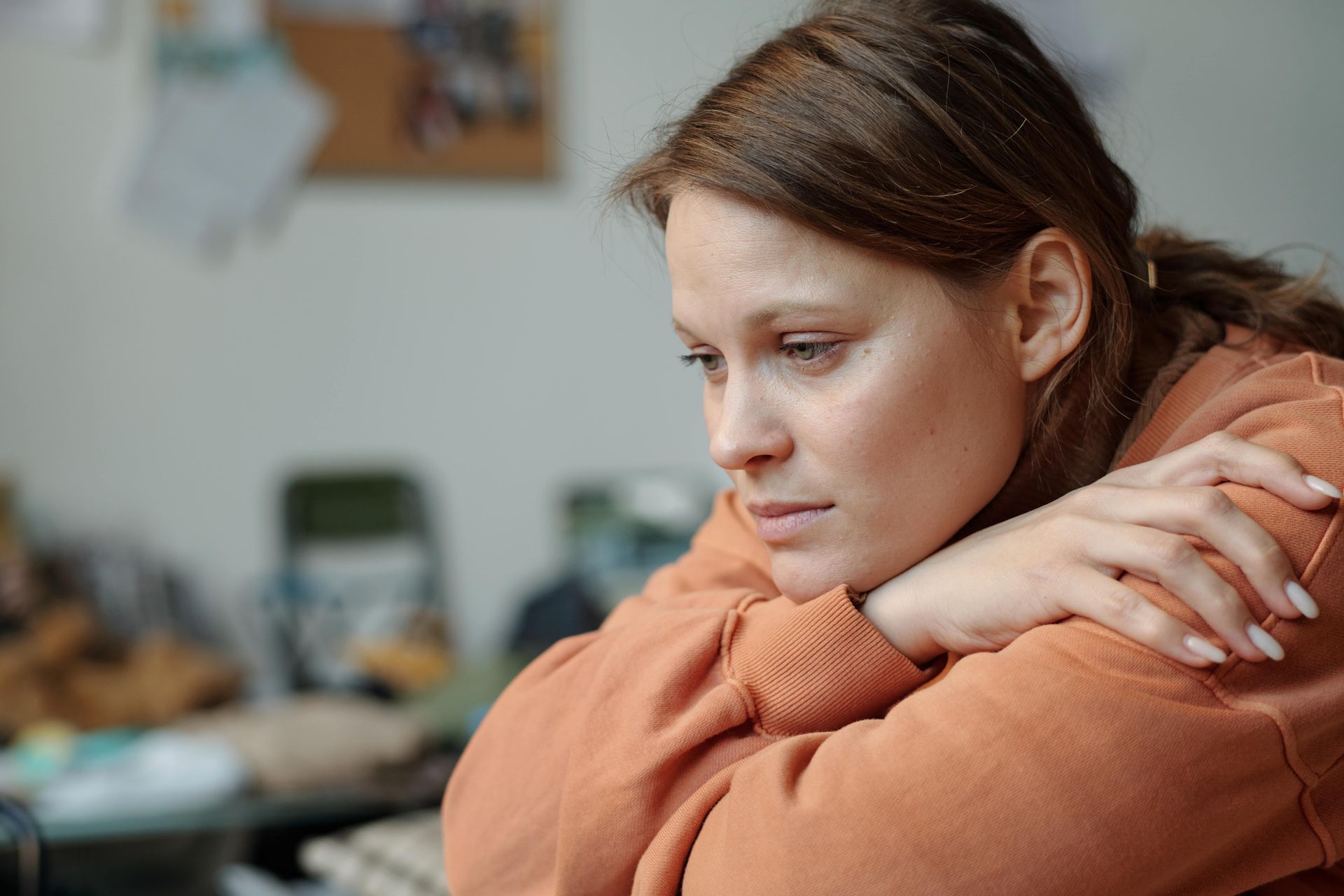 Woman in orange sweater, resting chin on arms, looking pensive in a room.