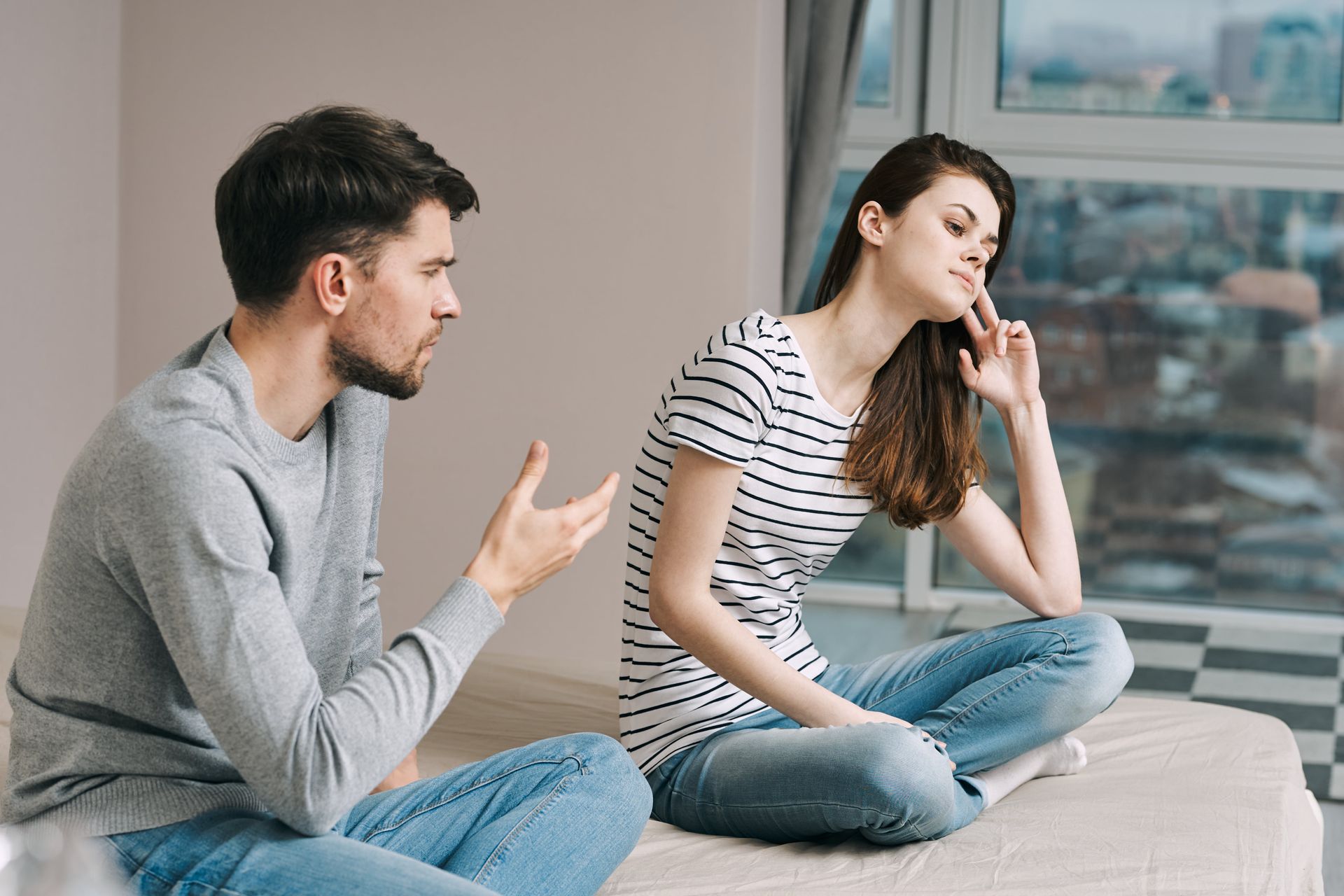 Man talking to woman sitting on bed, both look upset; modern bedroom setting.