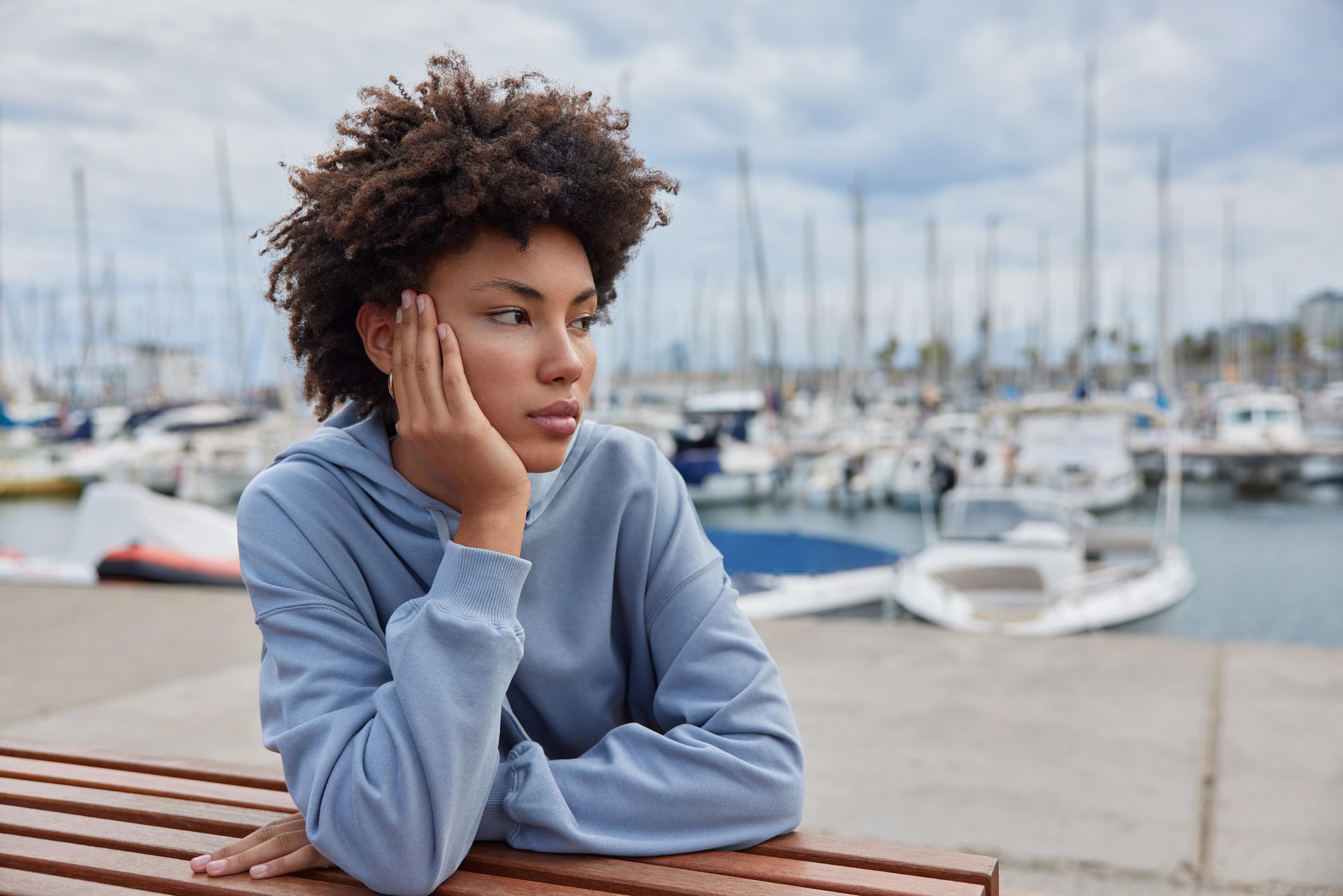 A young Black woman with curly hair looks pensive, resting her head on her hand, harbor in background.