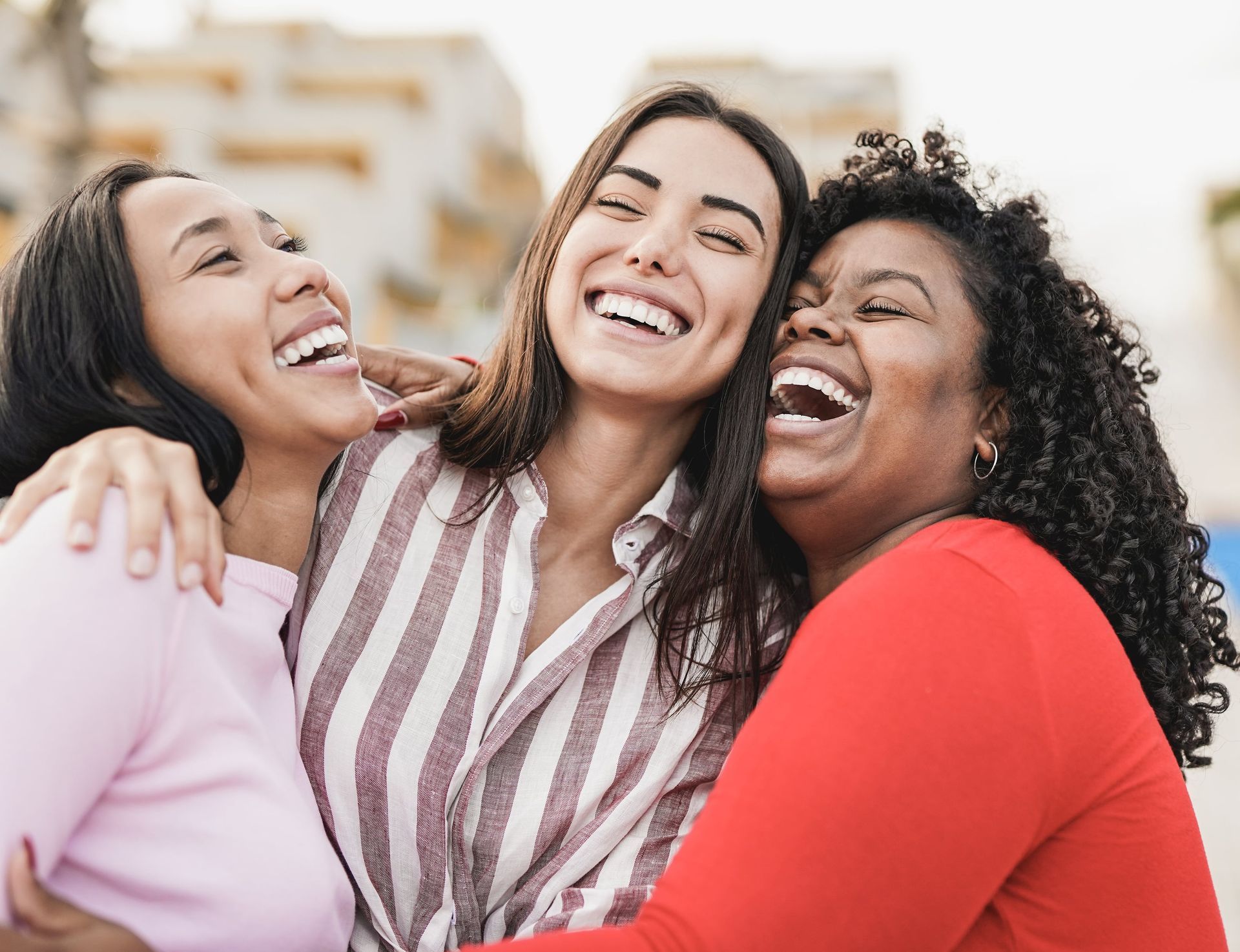 Three diverse women laughing, hugging outdoors.