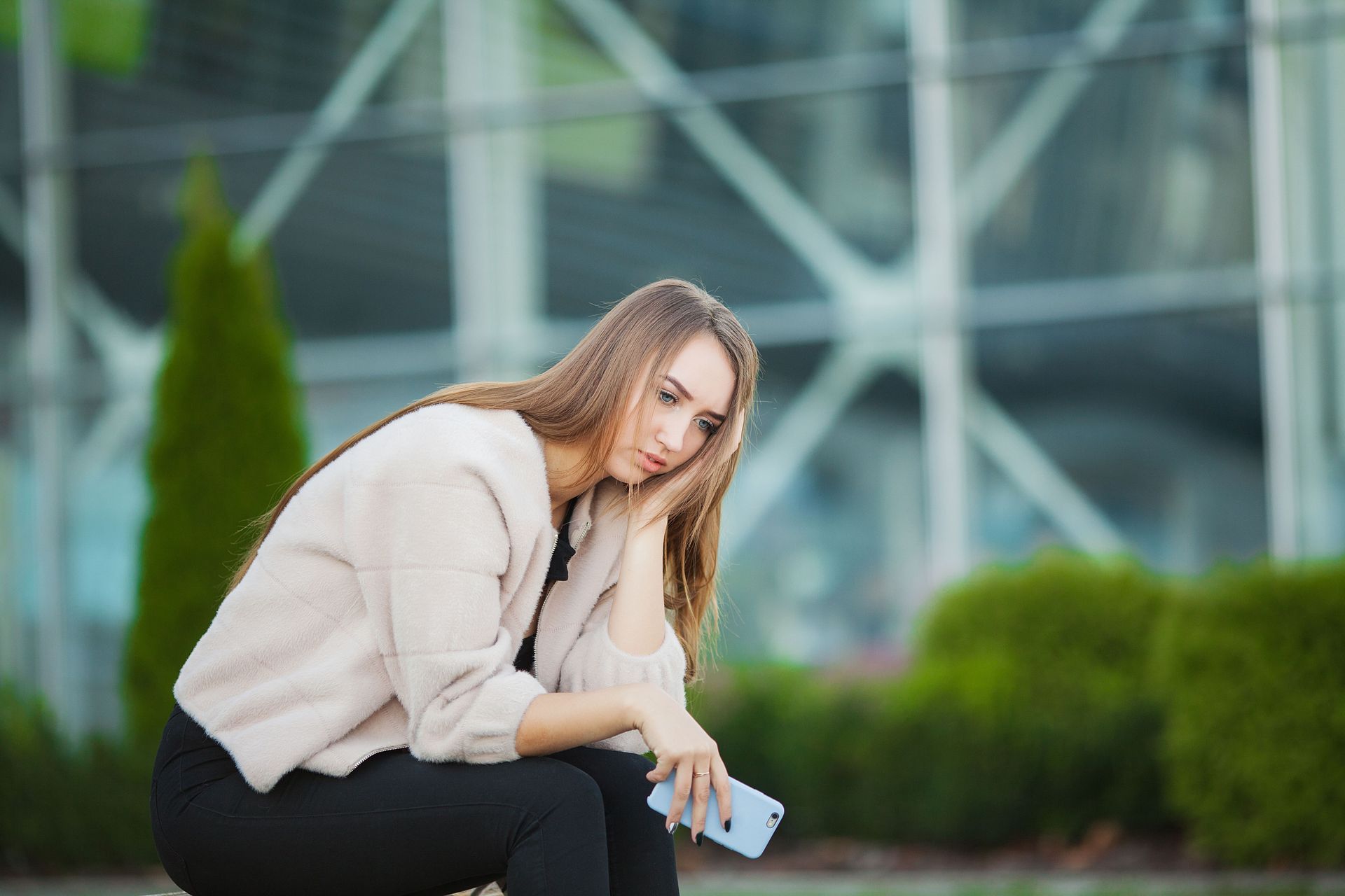 Woman in beige jacket sits outside, looking sad, holding a phone.
