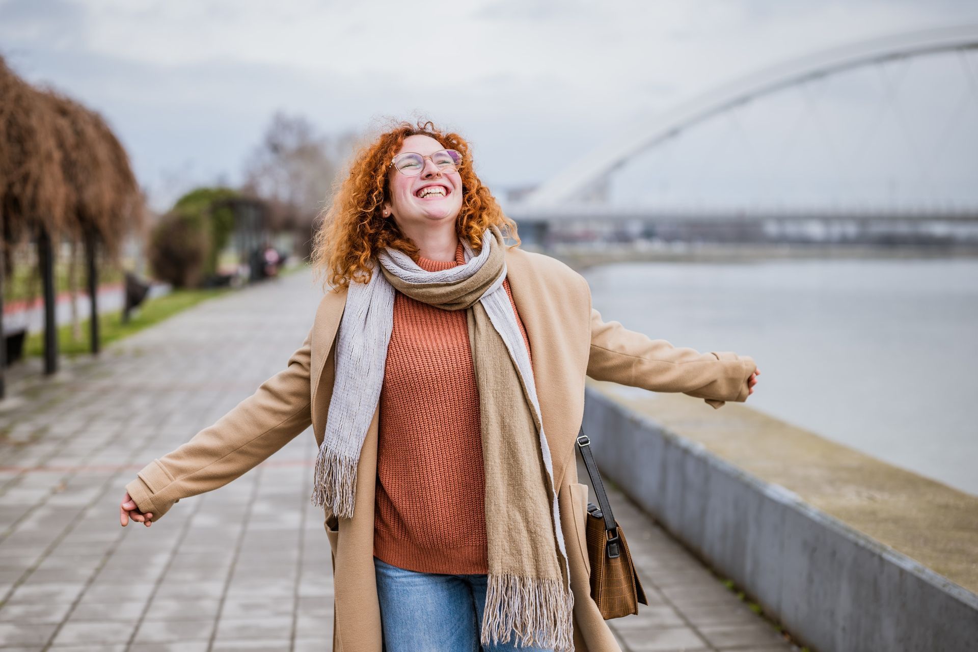 Woman with red curly hair smiles with arms outstretched on a waterfront promenade, bridge in background.