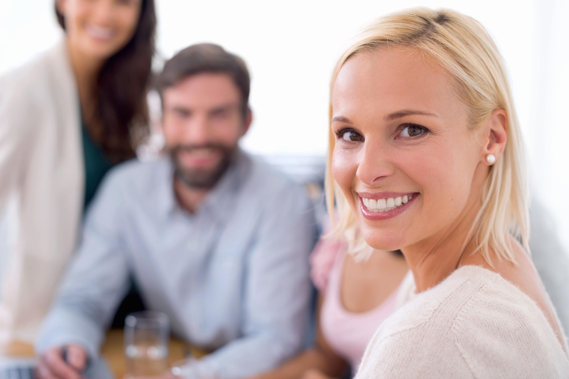 Woman with blonde hair smiles at the camera, surrounded by blurred coworkers in a light-filled office.