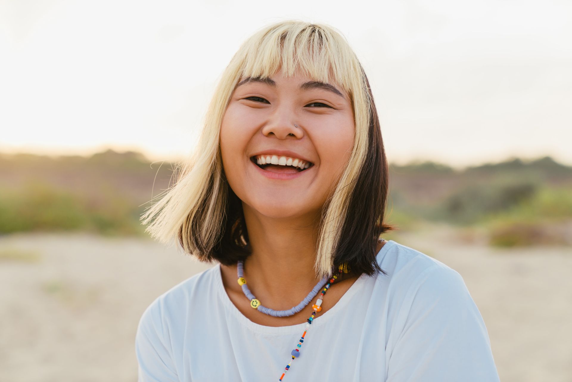Young woman with blonde bangs and a big smile, wearing a white shirt and beaded necklace, outdoors.
