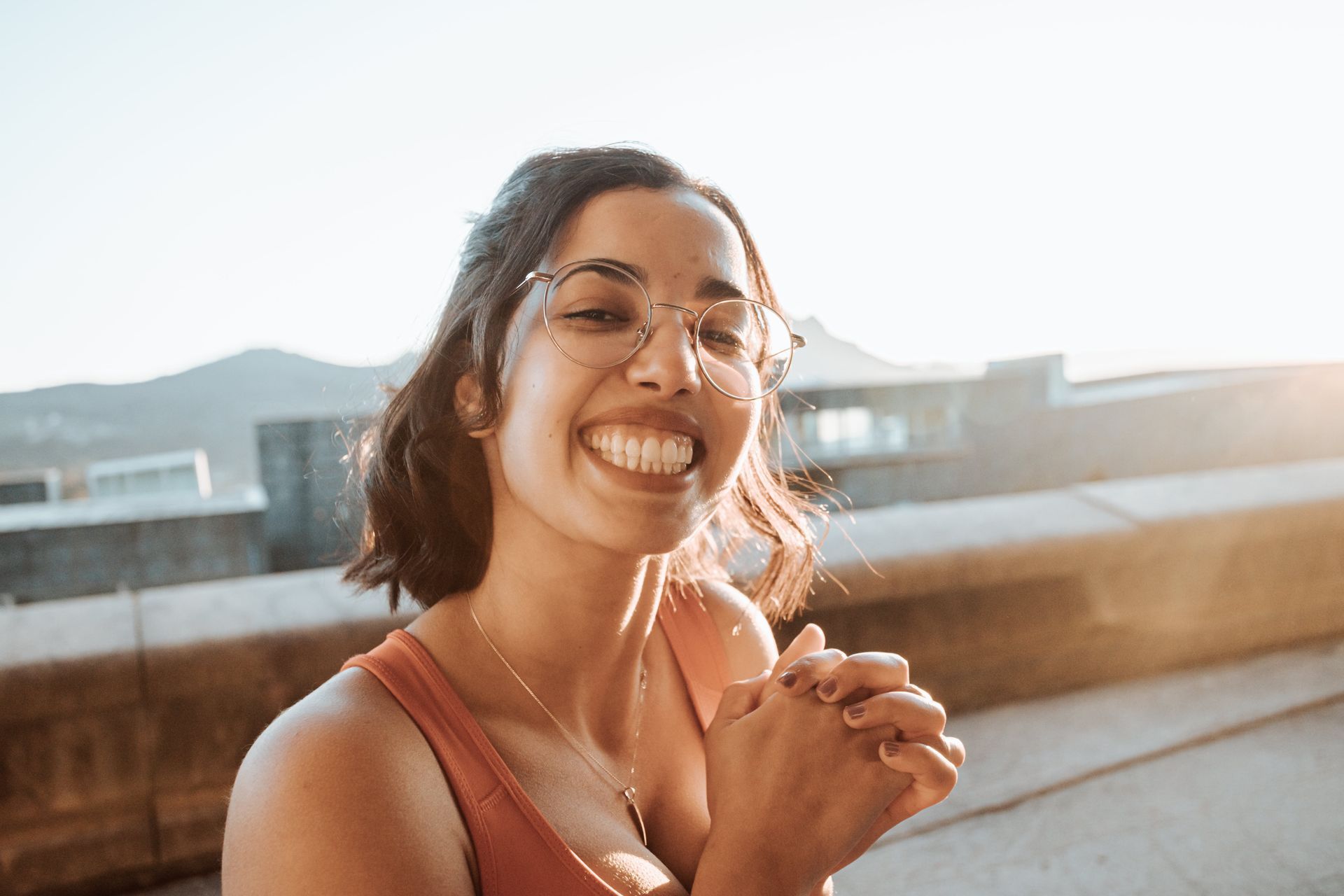 Woman with glasses smiles brightly outdoors.
