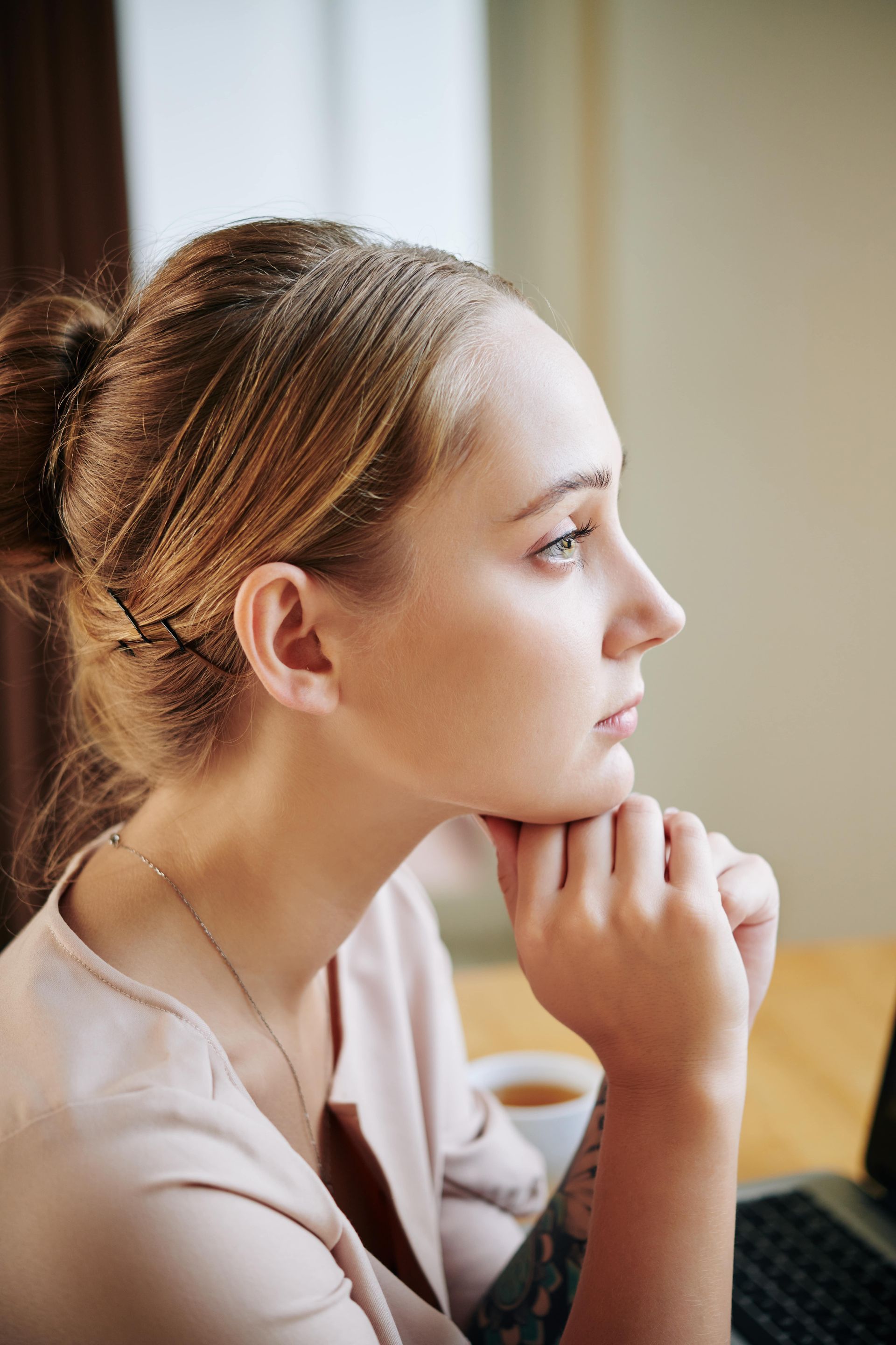 Woman with light brown hair in a bun, resting chin on hands, looking to the side in thought.