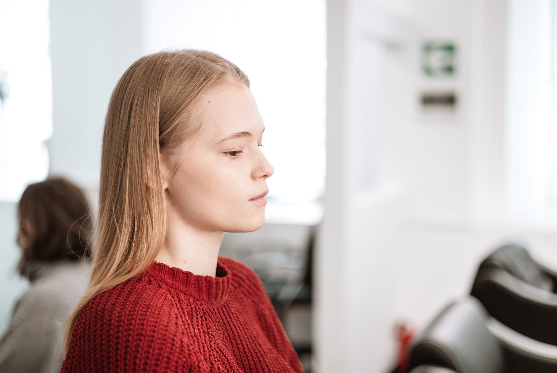 Woman with long blonde hair, wearing a red sweater, looks to the right in a white room.