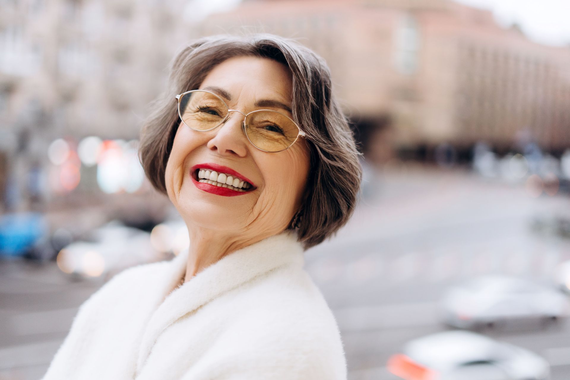 Smiling senior woman wearing glasses, light coat, red lipstick, outdoors in a city setting.