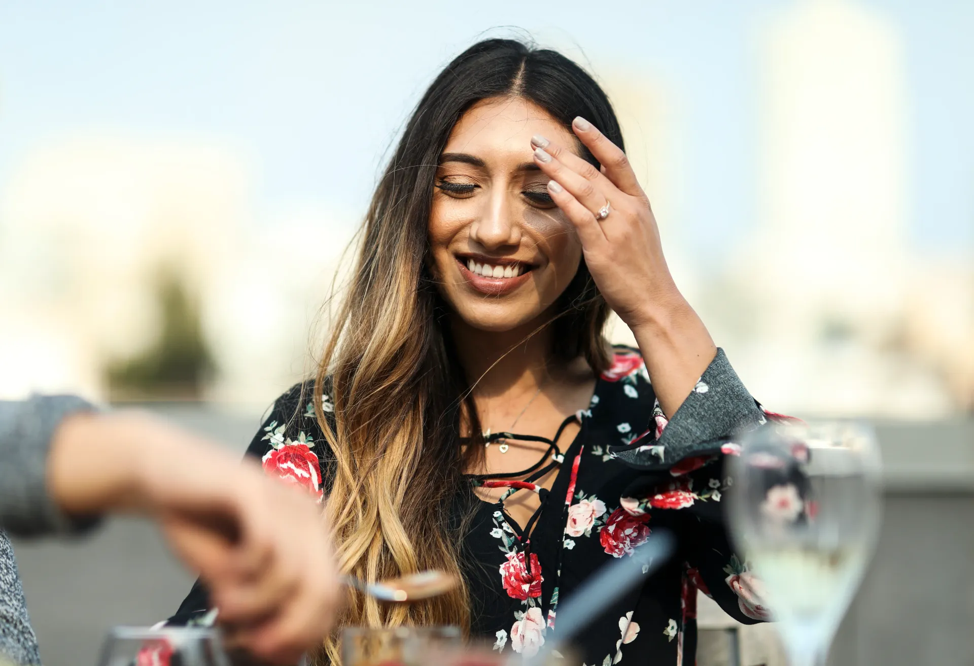 Woman smiles, touches hair while dining outdoors. She wears a floral shirt, sunny day.