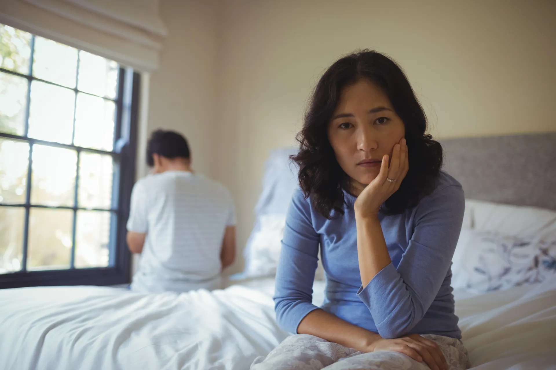 Woman looking at camera, hand on face, man sitting on bed, looking away; bedroom setting.