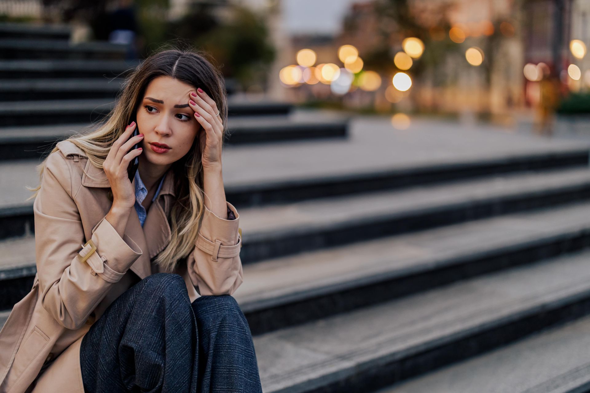 Woman in trench coat sitting on stairs, talking on phone, looking distressed.