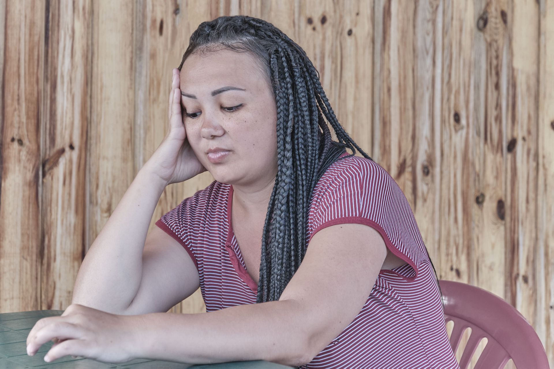 Woman with long braids resting her head on her hand, looking down, seated at a table, wooden backdrop.