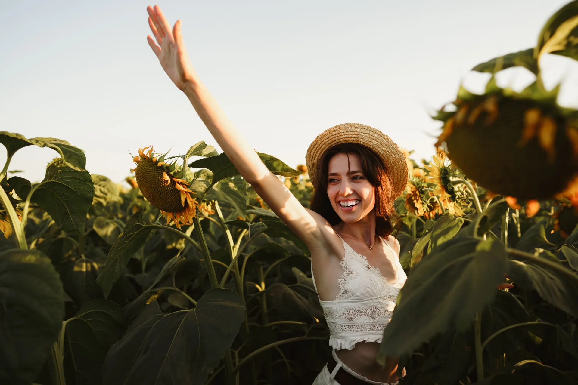 Woman in straw hat waving in a sunflower field, smiling.
