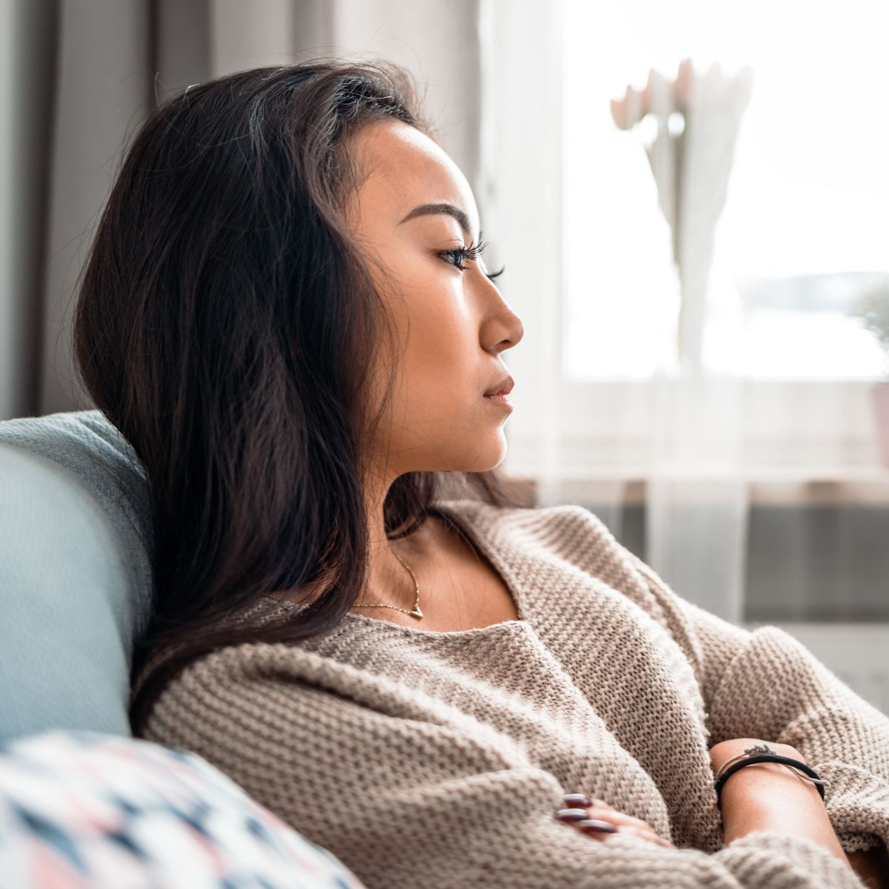 Woman with dark hair looks out window, resting on a couch, wearing a neutral-colored sweater.