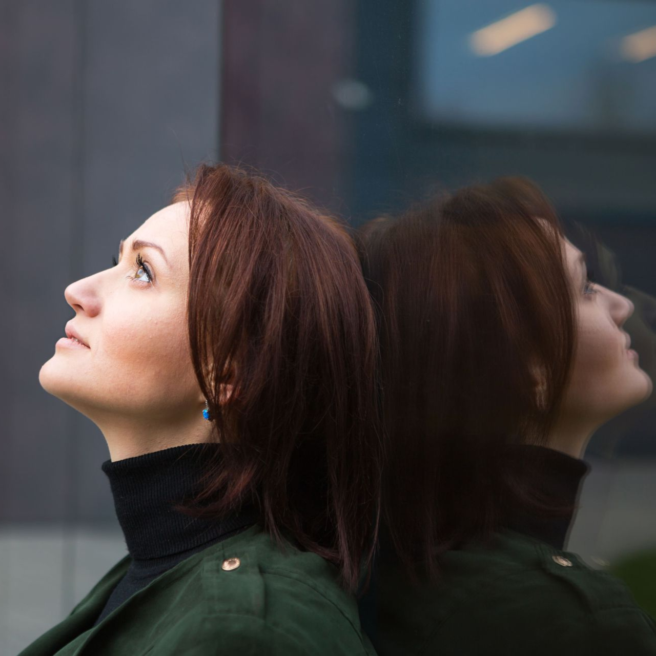 Woman with auburn hair looks up, leaning against reflective glass. Dark turtleneck, green coat.