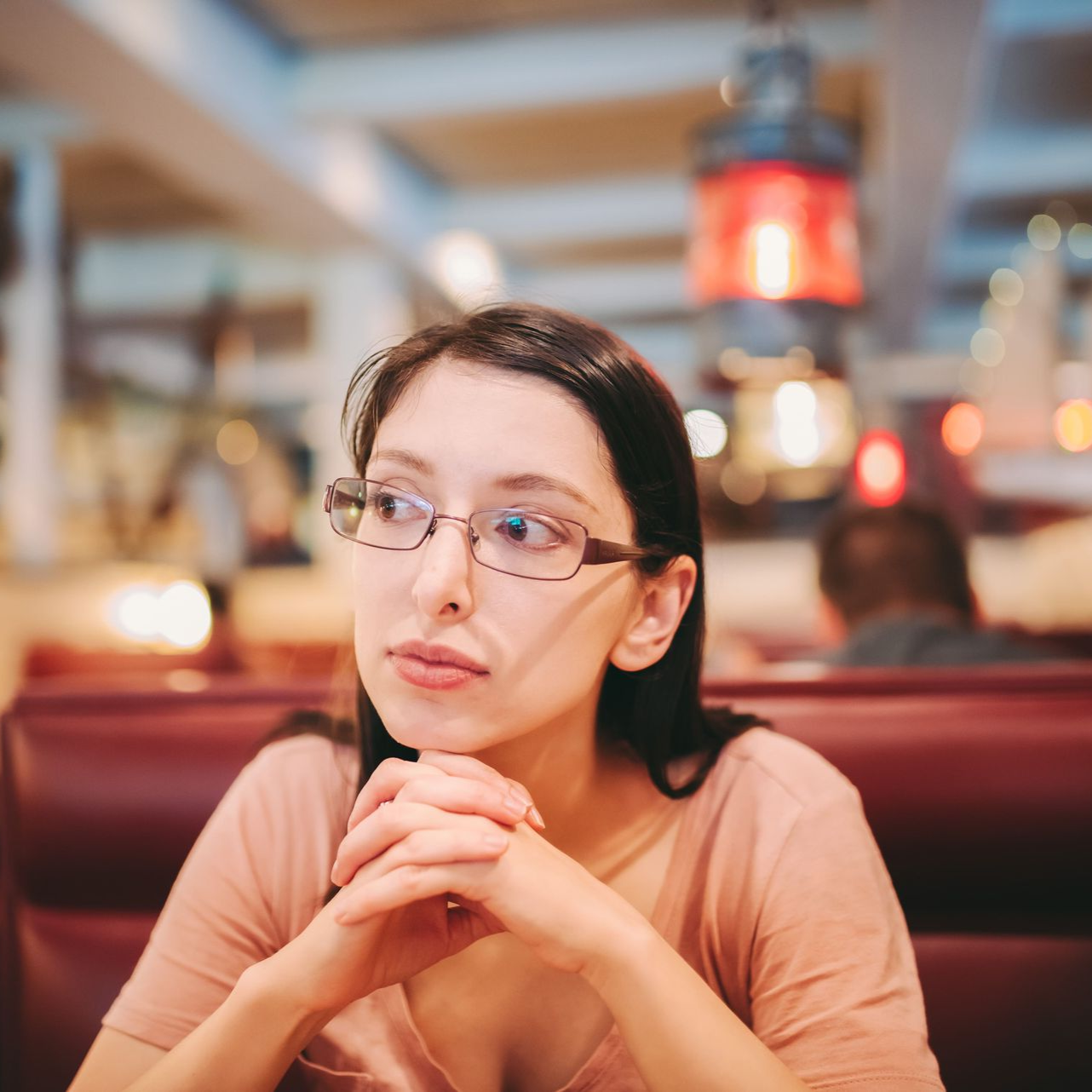 Woman in glasses at a restaurant, hands clasped, looking thoughtful. Pink shirt, booths, blurred background, red light.
