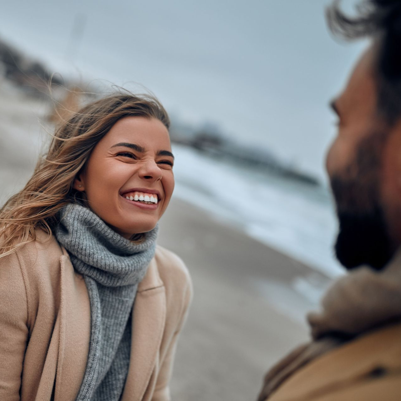Woman laughing, looking at a man on a beach. She wears a tan coat and scarf; cloudy sky in the background.