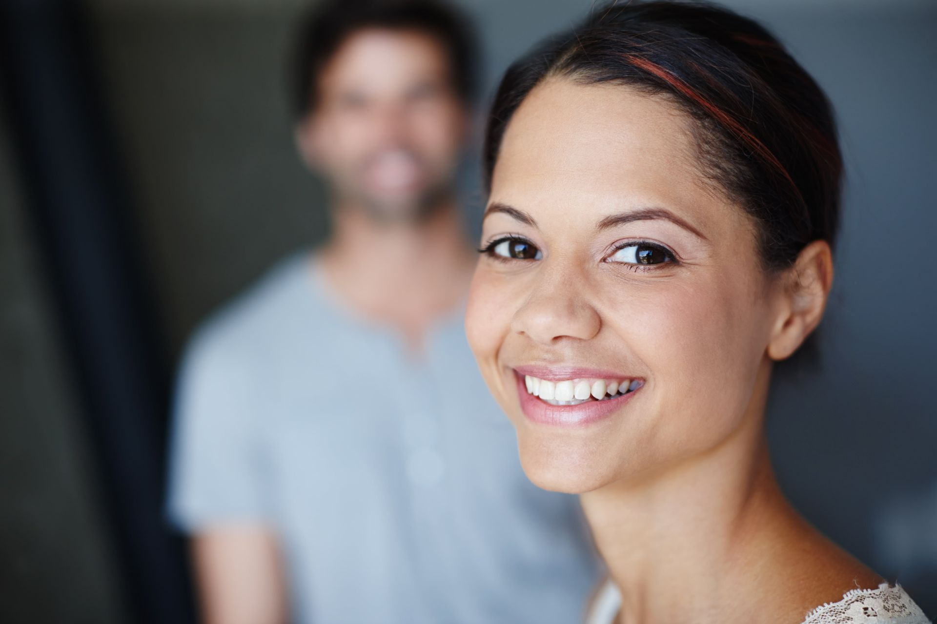 Woman smiling, man blurred in background. Indoor, natural light.