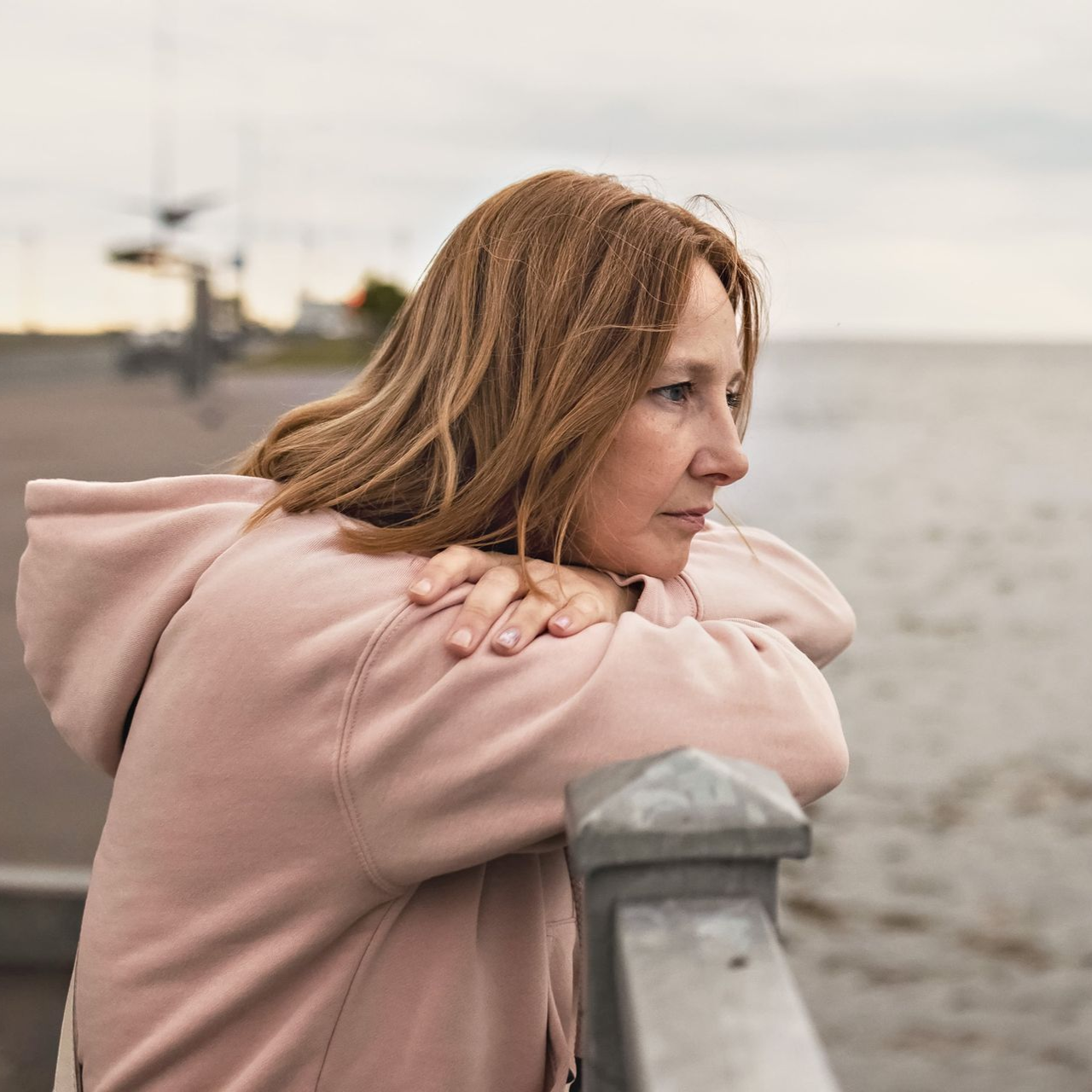 Woman in pink hoodie leaning on railing, looking out at water.