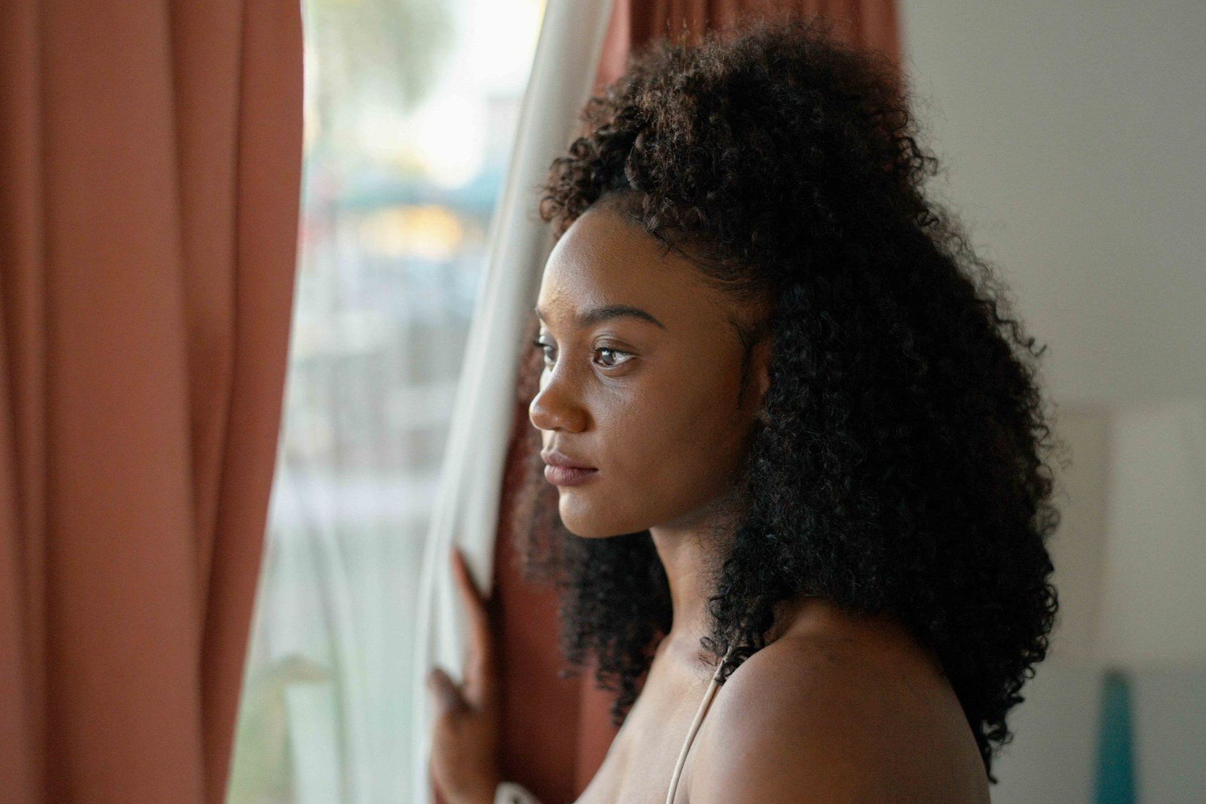 Woman with curly brown hair, wearing a brown sweater, gazes thoughtfully to the side. Sitting in a chair near a window.