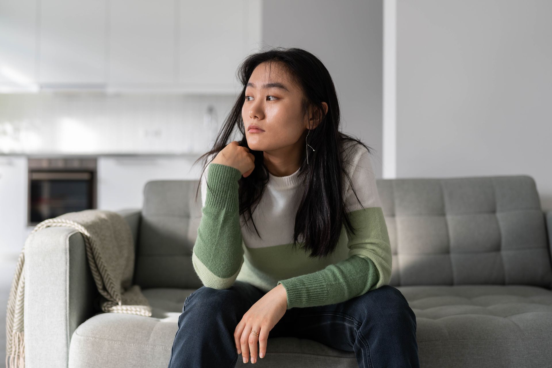 Woman on a gray sofa, looking away with a pensive expression. She wears a green and white top and jeans. Indoors.