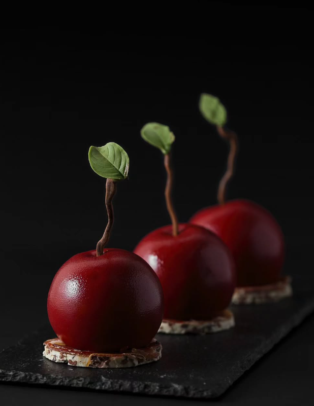 Three red, glossy dessert spheres resembling apples with chocolate stems and green leaves, on round crackers, black background.