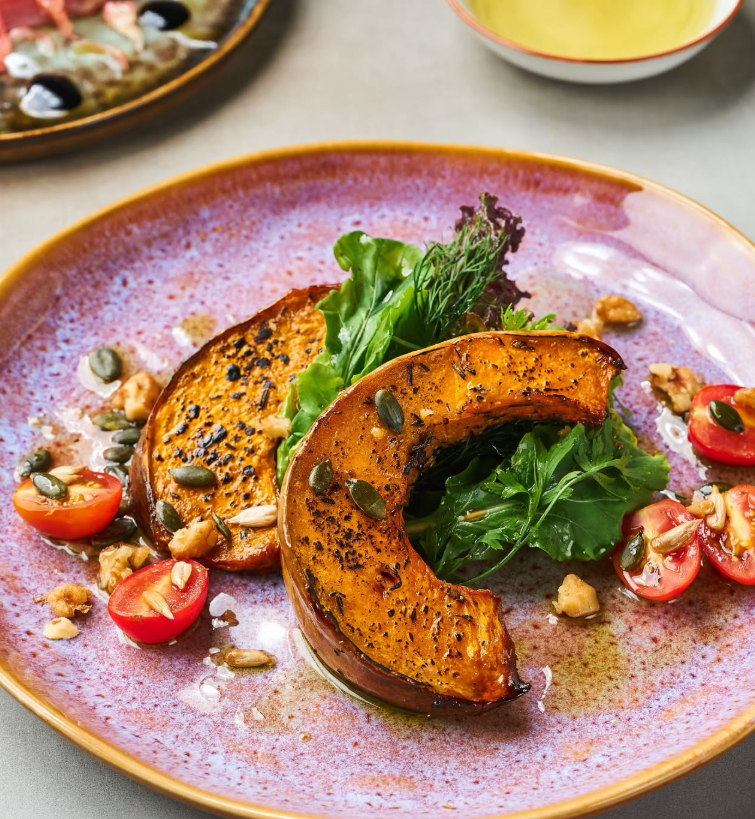 Roasted pumpkin with greens, tomatoes, nuts, and seeds on a pink plate.