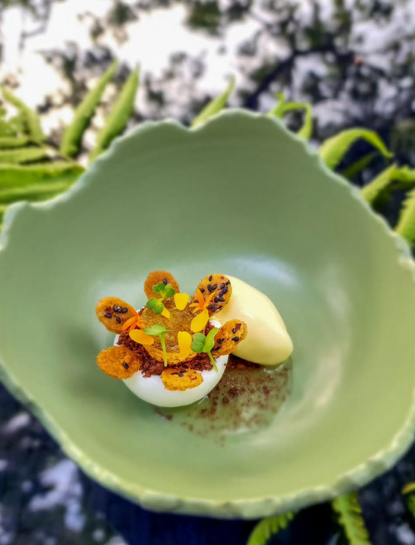 A half-egg dish with a topping of flowers and a scoop of pale yellow cream in a leaf-shaped green bowl.