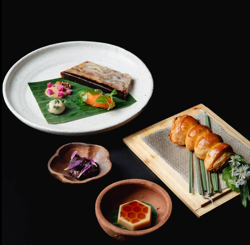 Deconstructed meal with various small dishes: meat, vegetables, rolls, honeycomb, all on black surface.