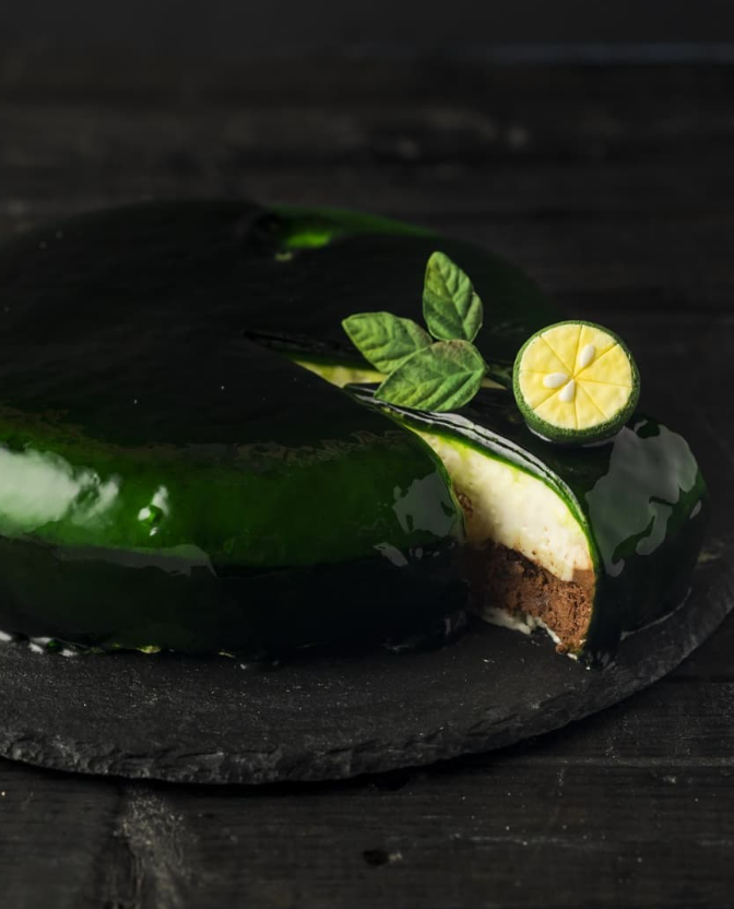 Green glazed cake with a slice removed, garnished with lime and mint on a dark surface.