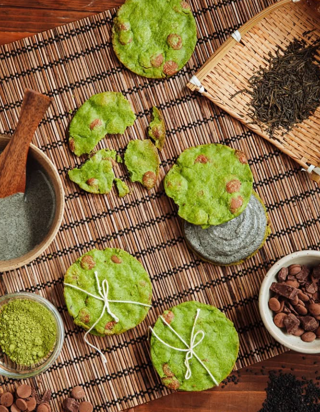 Green matcha cookies with chocolate chips, set with matcha powder, tea leaves, and wooden tools.