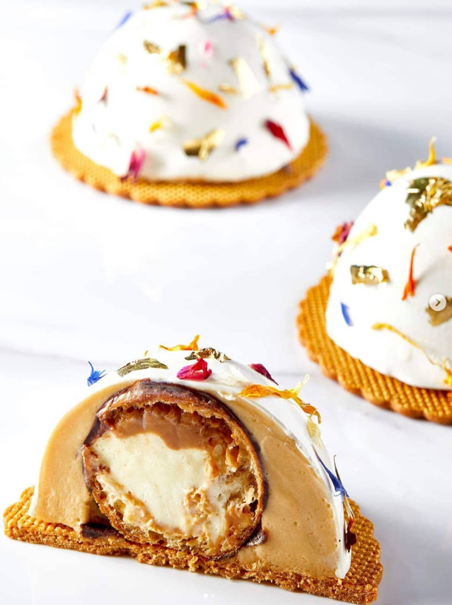 Three dome-shaped desserts: white cream topped with edible flowers and gold leaf, set on a cookie base; one is cut open.