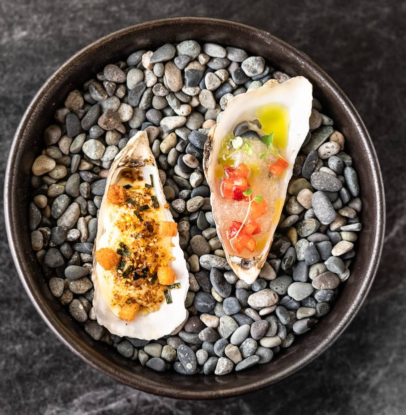 Two oysters, one baked with topping, one raw with tomato, in a bowl on pebbles.