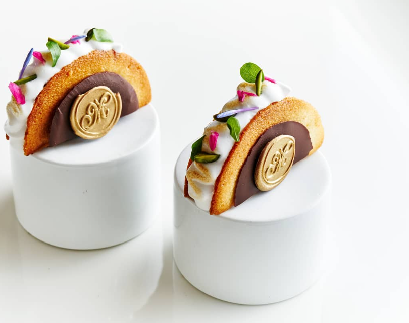 Two dessert tacos on white pedestals: curved pastry shell filled with white cream and topped with greens, chocolate coin.