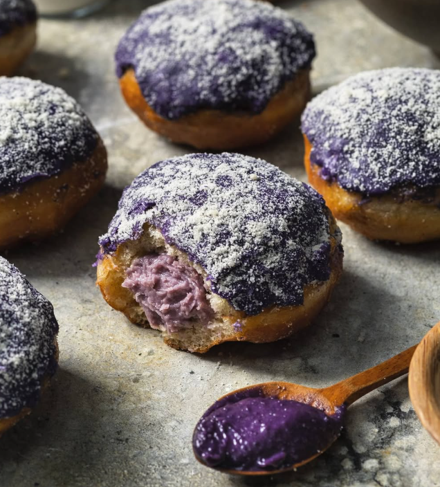 Ube-filled donuts with purple frosting, one bitten into, on a gray surface, a spoonful of filling.