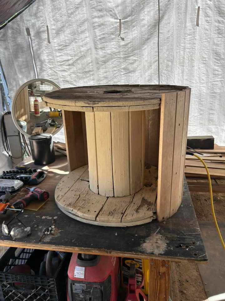 Wooden cable spool repurposed as a table on a workbench. Light wood, some tools, outdoors.