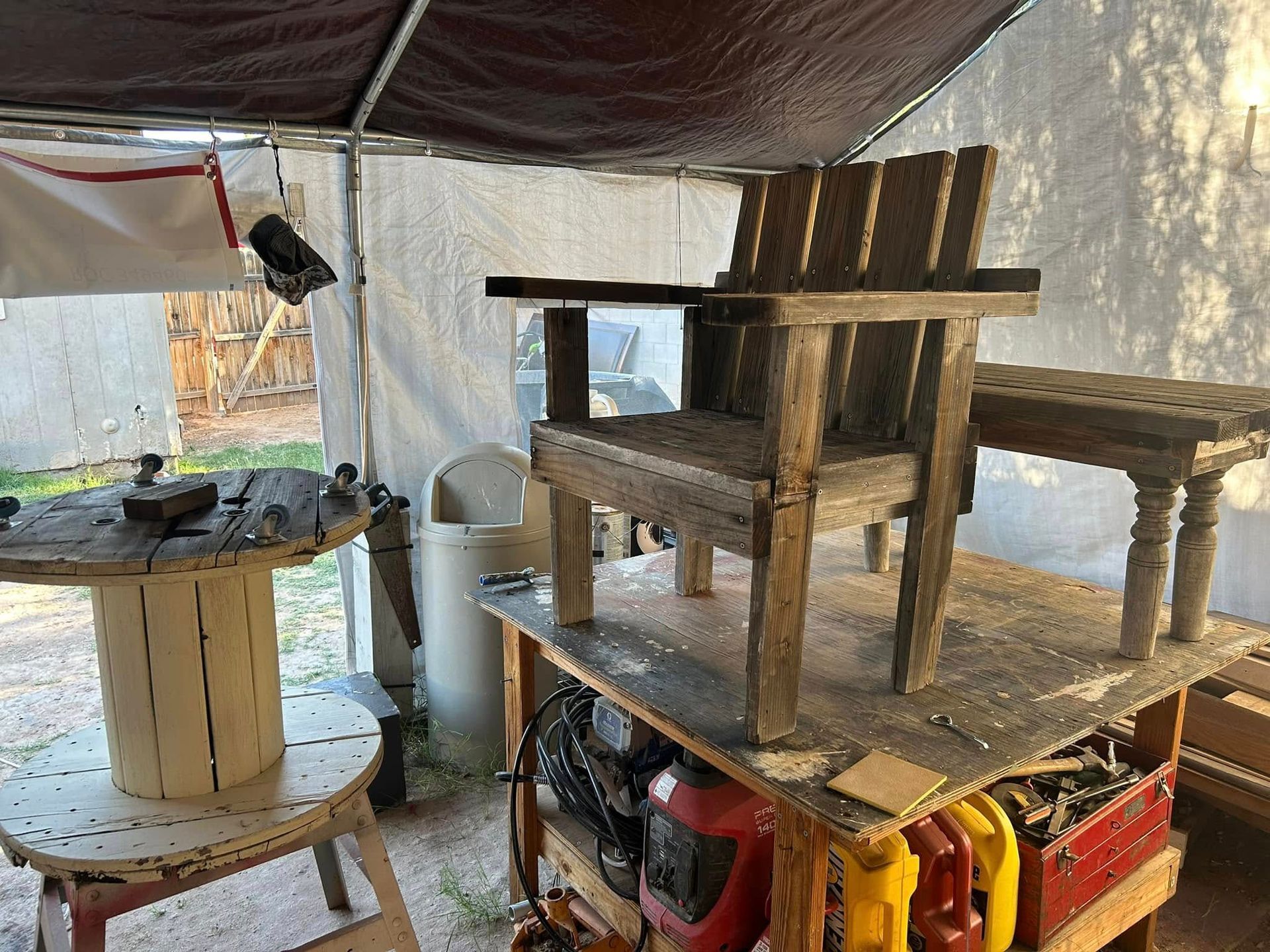 Rustic wooden chair and small table on a workbench under a tarp; other tools and materials visible.