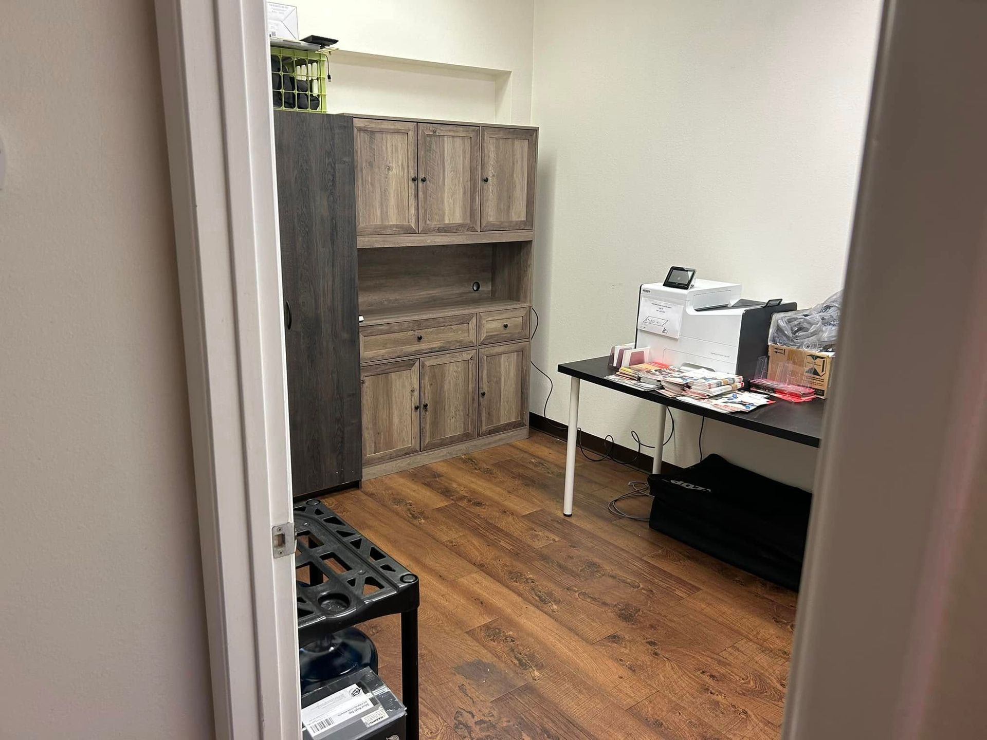 Small office with wooden cabinets, a desk, and wood flooring, viewed through an open doorway.