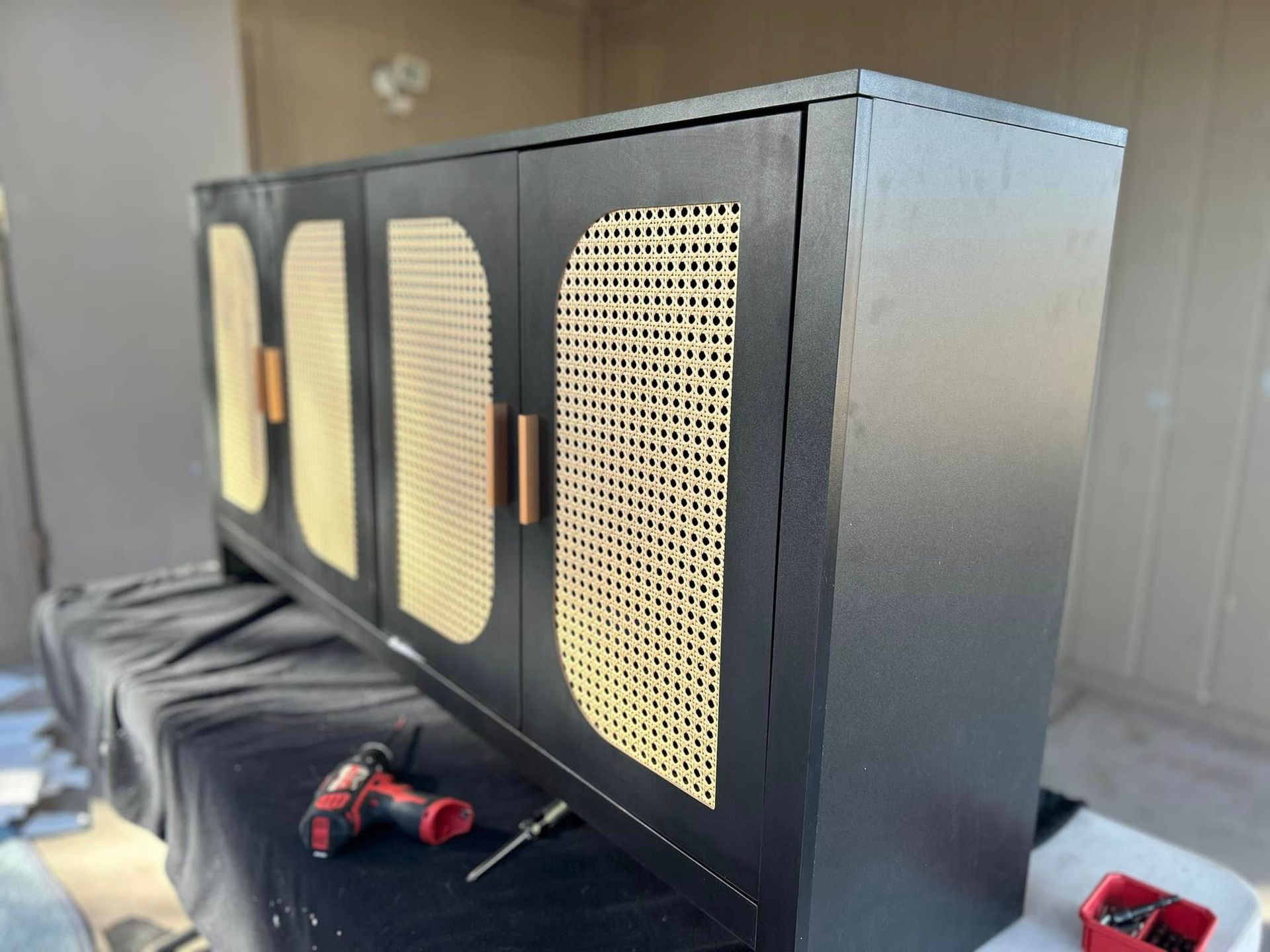 Black cabinet with woven cane door panels, copper handles, resting on a table with tools.